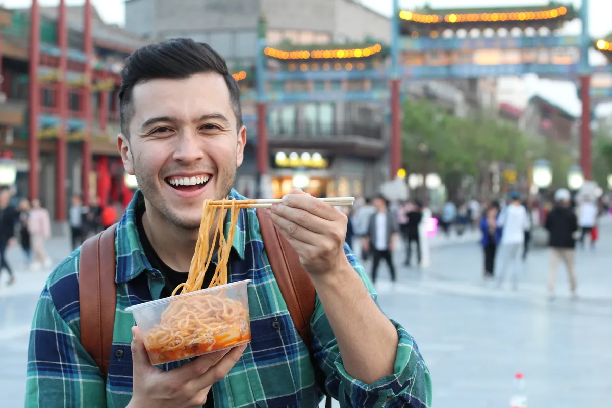 A smiling traveler eating noodles with chopsticks on a Beijing street lined with red gateways