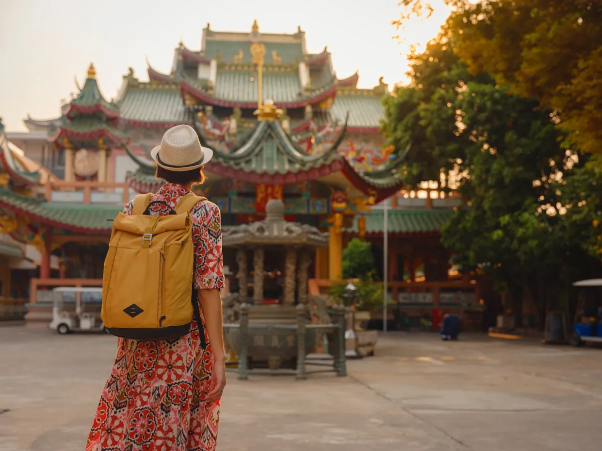 A traveler with a yellow backpack walking toward an ornate Chinese temple with green tiled roofs