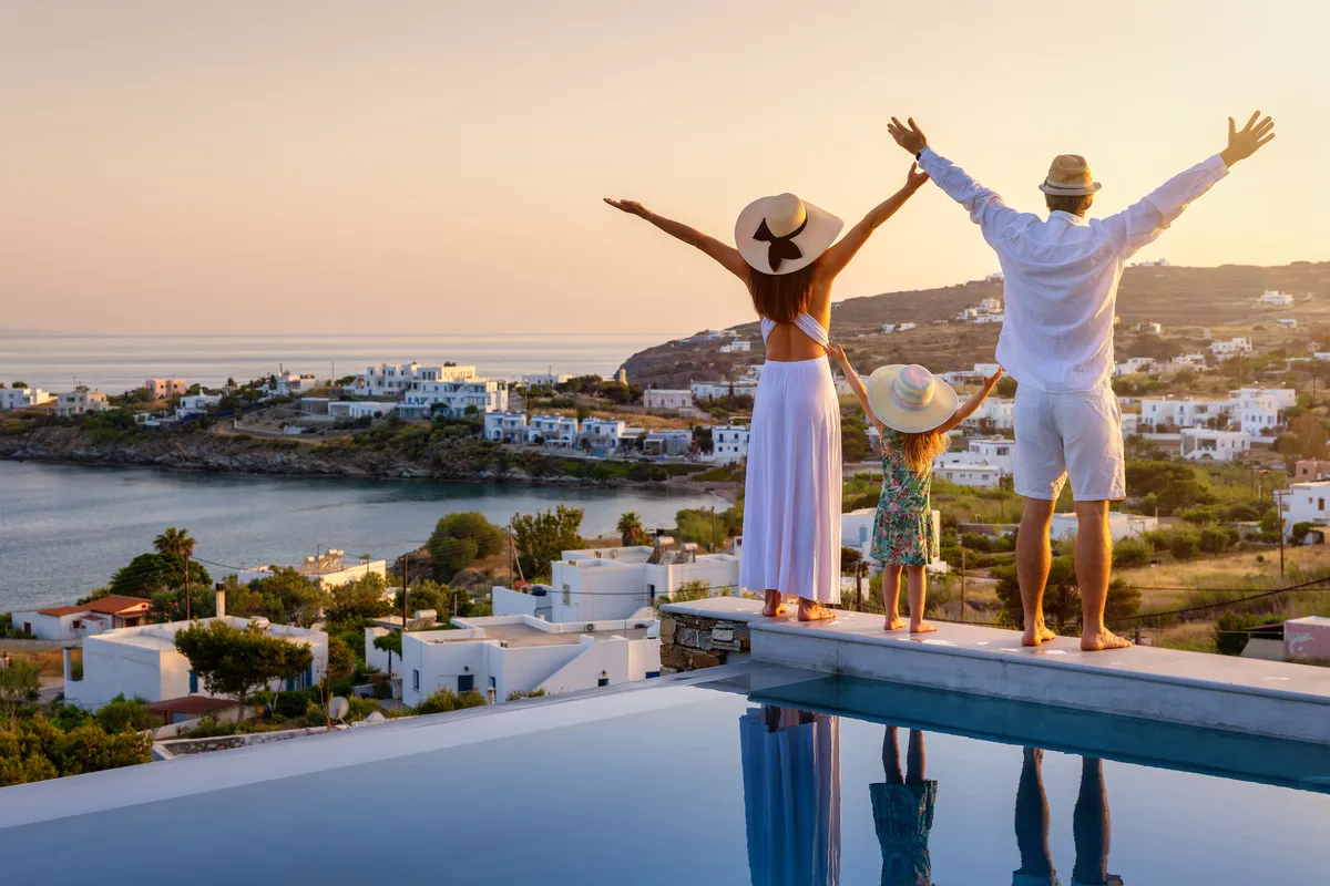 A family with arms raised on a pool terrace above the whitewashed village of a Greek island at sunset